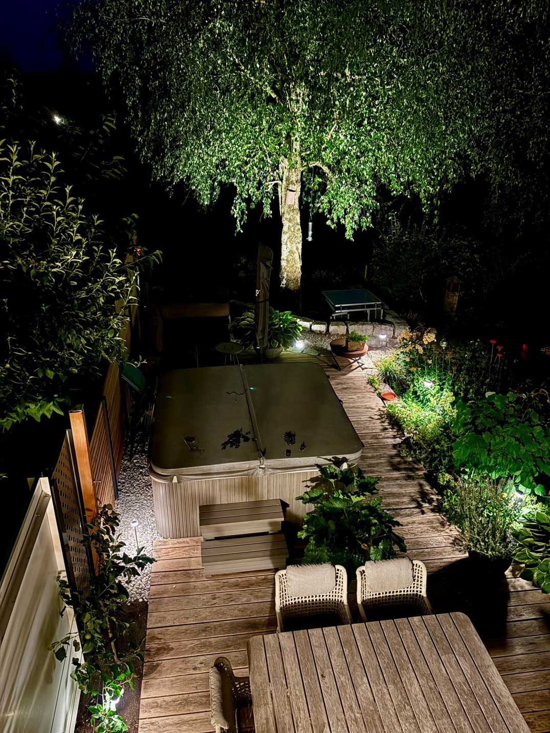 Stimmungsvolle Gartenbeleuchtung mit beleuchtetem Baum, Holzterrasse, Pflanzenbeeten und Whirlpool im privaten Garten